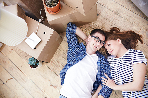 Lovely young couple lying on wooden floor of their new apartment and sharing  ideas concerning interior design of living room, pile of moving boxes standing next to them