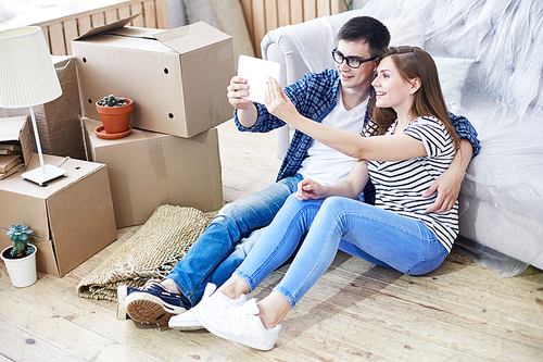 Handsome young man and his beautiful girlfriend sitting on floor of new apartment and taking selfie on digital tablet, pile of moving boxes and interior design items on background