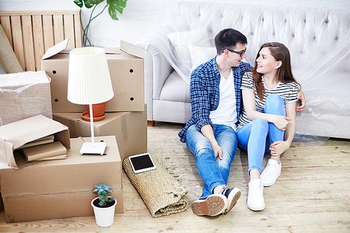 Lovely young couple wearing jeans and T-shirts chatting animatedly with each other while sitting on floor of new apartment, pile of moving boxes and interior design items on background