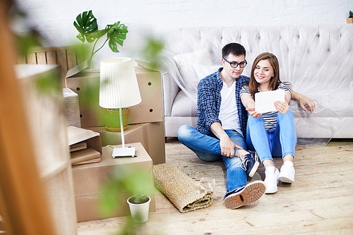 Full length portrait of lovely young couple wearing jeans and T-shirts taking selfie with help of digital tablet while sitting on wooden floor of new apartment, piles of moving boxes on background