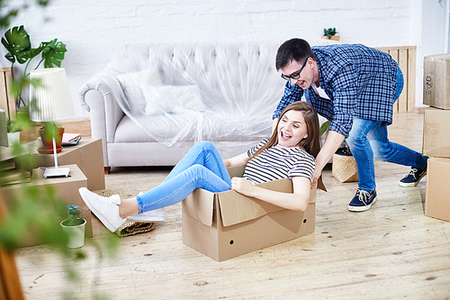 Lovely young couple having fun while riding in cardboard box at new apartment, interior design items and moving boxes on background