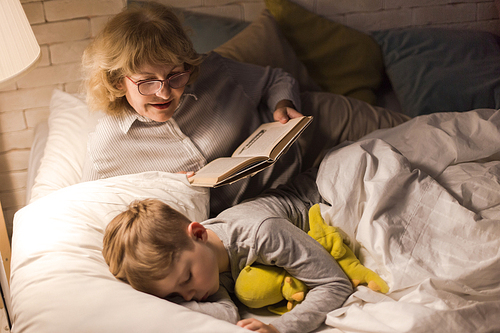 Portrait of smiling grandmother wearing glasses reading bedtime stories to sleeping little boy in dark room under lamplight
