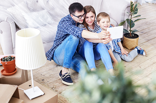 Adorable family of three gathered together in living room of new apartment and taking selfie on digital tablet, interior design items and moving boxes on background