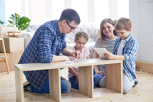 Loving young family of four gathered together at living room of new apartment and wrapped up in assembling wardrobe, pile of moving boxes on background