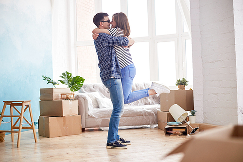 Profile view of loving young couple expressing their feeling while standing at living room of new apartment, interior design items and moving boxes on background