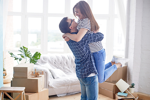 Cheerful young man wearing checked shirt and jeans holding his pretty girlfriend in arms while standing at living room of new apartment,  interior design items and moving boxes on background