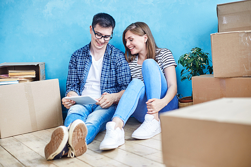 Full length portrait of loving young couple wearing jeans and sneakers looking through funny photos on digital tablet while sitting on wooden floor of new apartment.