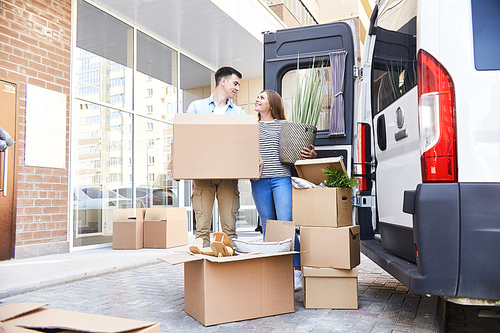 Portrait of happy young couple posing with cardboard boxes standing next to moving van outdoors