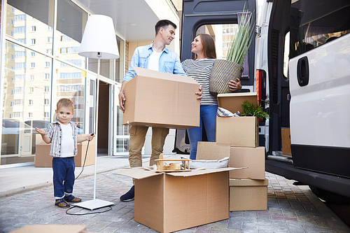 Portrait of happy young family holding cardboard boxes standing next to moving van and smiling