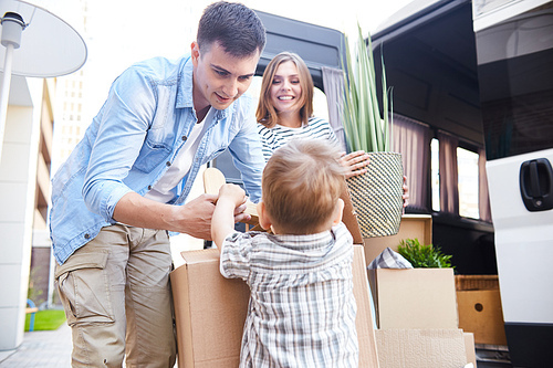 Portrait of happy young family loading boxes to moving van, little boy helping father pack his toys