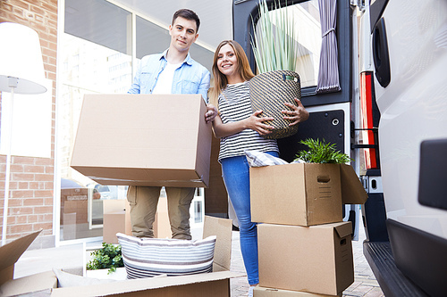 Portrait of happy young family posing with cardboard boxes and houseplant standing next to moving van outdoors smiling at camera