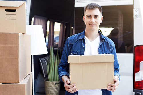 Portrait of smiling young man holding cardboard box and  while unloading moving van outdoors