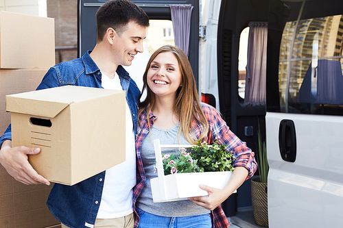 Portrait of happy  young couple embracing and smiling at camera holding cardboard boxes next to moving van outdoors