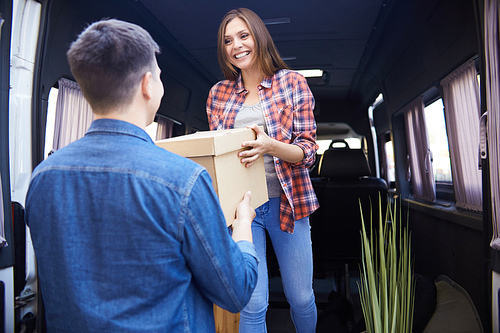 Portrait of young happy woman unloading cardboard boxes from moving van handing them to husband