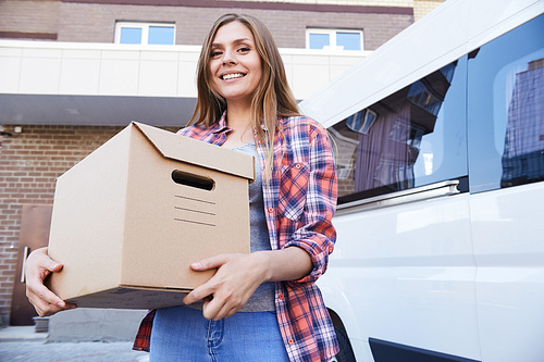 Portrait of happy young woman holding cardboard box and smiling to camera posing next to moving van