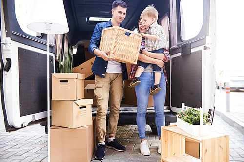 Portrait of happy young family moving into new home, standing by moving van holding boxes outdoors