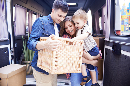 Portrait of happy young family with little boy unloading moving van outdoors and unpacking boxes smiling brightly