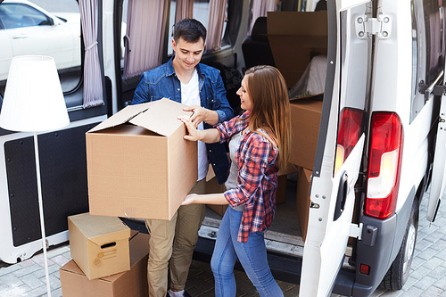 Portrait of smiling young man and woman unloading big cardboard boxes from moving van outdoors