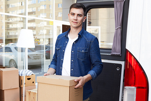 Portrait of young man holding cardboard box and smiling happily to camera  while unloading moving van outdoors