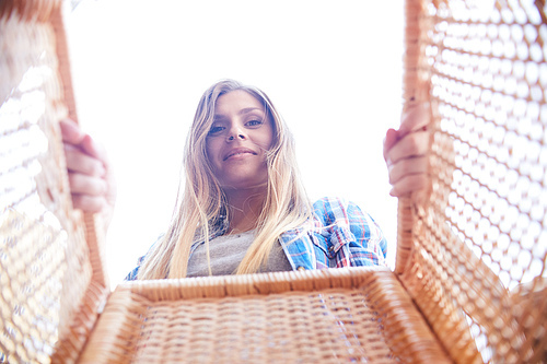 Portrait of happy young woman looking into wicker basket and smiling, shot from inside the box against white sky