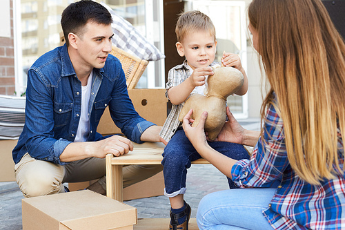 Portrait of happy young family unpacking cardboard boxes outdoors while moving in little boy helping  pack his toys