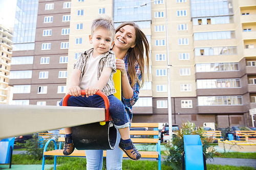 Portrait of young mother playing with cute little boy outdoors, swinging on modern playground in front of apartment building