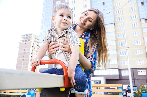 Portrait of young mother playing with little boy outdoors, swinging on modern playground in front of apartment building