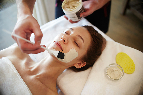 Portrait of beautiful young woman in SPA, lying on massage table enjoying face masks and beauty treatments, with male hands applying clay cream