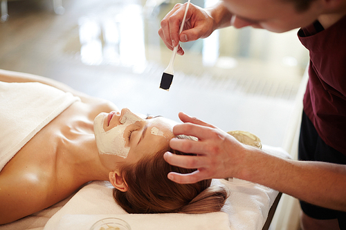 Side view of unrecognizable cosmetologist applying face mask with brush to face of beautiful woman lying on massage table in SPA center