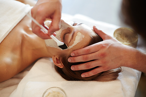 Closeup of unrecognizable cosmetologist applying face mask with brush to face of beautiful woman lying on massage table in SPA center