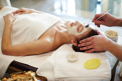 Side view portrait of beautiful young woman in SPA, lying on massage table with cosmetologist applying face mask to her skin