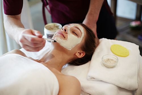 Closeup of unrecognizable male cosmetologist applying face mask with brush to face of beautiful woman lying on massage table in SPA center