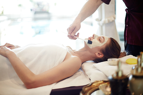 Side view portrait of beautiful young woman in SPA, lying on massage table with eyes closed while cosmetologist applying face mask to her skin