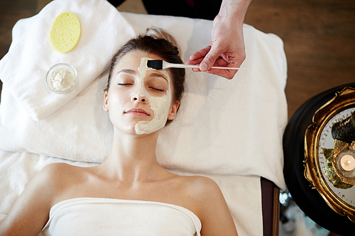 Top view portrait of beautiful young woman in SPA, lying on massage table with cosmetologist applying face mask to her face