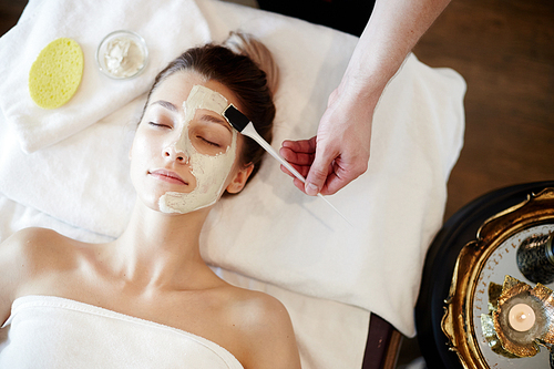 Top view portrait of beautiful young woman enjoying SPA, lying on massage table with cosmetologist applying face mask to her face with brush