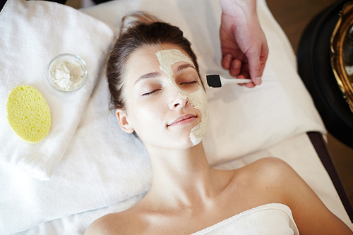 Top view portrait of beautiful young woman in SPA, lying on massage table with eyes closed while cosmetologist applying face mask to her face