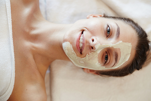 Top view closeup portrait of blissful young woman enjoying beauty treatments and face masks in SPA, lying on massage table smiling happily at camera