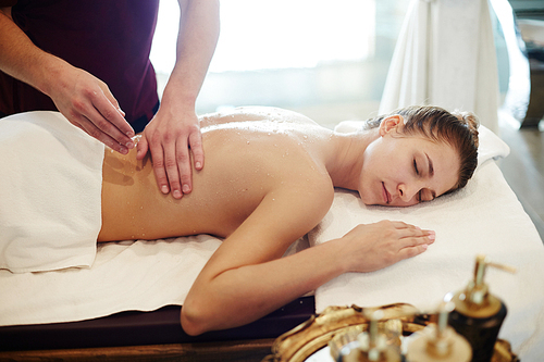 Portrait of blissful young woman enjoying massage in SPA center, lying on massage table with eyes closed and smiling with male masseur rubbing her back