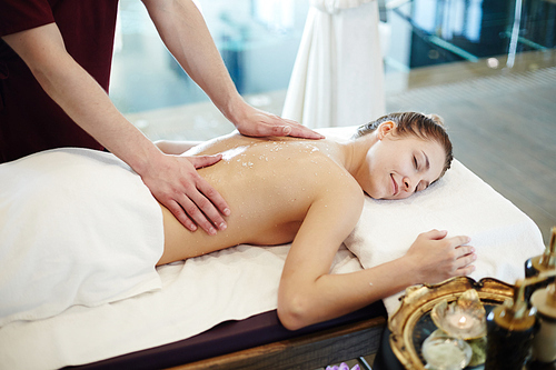 Portrait of blissful young woman enjoying massage in SPA center, lying on massage table with eyes closed and smiling