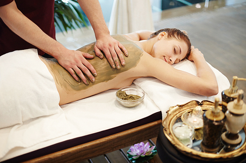 Portrait of blissful young woman enjoying mud massage in SPA center, lying on massage table with eyes closed and smiling