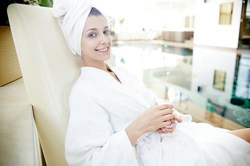 Portrait of beautiful young woman smiling at camera relaxing in lounge chair by swimming pool wearing bath robe