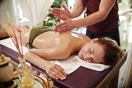 Portrait of beautiful young woman lying on massage table with eyes closed and smiling blissfully enjoying SPA treatment, man massaging her with lotions and body oils