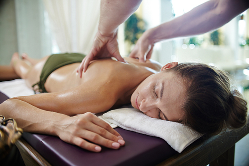 Portrait of  young woman lying on massage table with eyes closed  blissfully enjoying SPA treatment, man massaging her with lotions and body oils