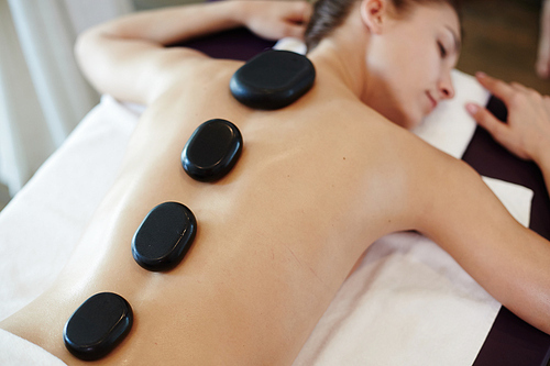 Above view portrait of young woman lying on massage table in SPA center with hot stones in row on her back