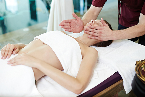 Portrait of beautiful young woman lying on massage table in SPA center with man massaging her body and face