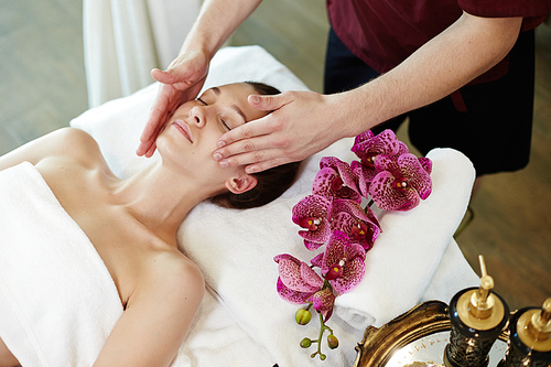 Portrait of beautiful young woman enjoying face lifting massage lying on table in SPA center and relaxing with male masseuse rubbing her skin