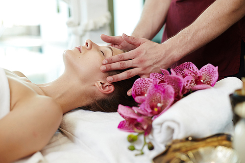 Side view portrait of beautiful young woman enjoying face lifting massage lying on table in SPA center and relaxing with male masseuse rubbing her temples