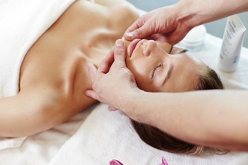 Portrait of beautiful young woman lying on massage table in SPA with eyes closed enjoying face massage