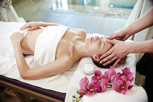 High angle portrait of young woman enjoying face and head massage lying on table in SPA center and relaxing