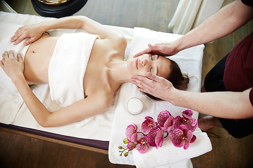 High angle portrait of young woman enjoying face lifting massage lying on table in SPA center and relaxing, smiling blissfully